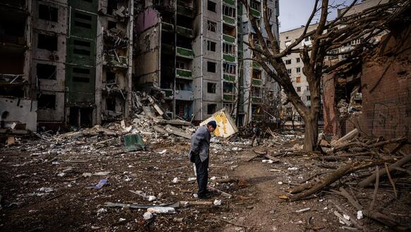 Das Bild zeigt eine Person, die vor einem stark zerstörten Haus steht