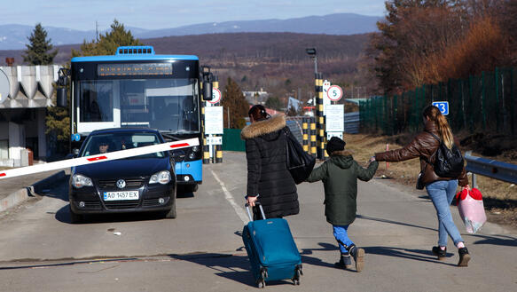 Das Bild zeigt zwei Erwachsene Menschen, die die Hände eines Kindes halten und zusammen mit Gepäck in Richtung eines Busses hinter einer Schranke laufen.
