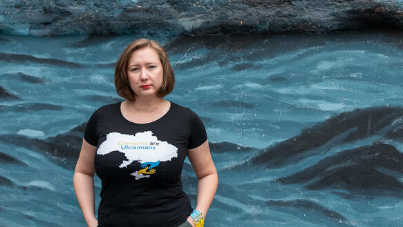 Eine Frau mit kinnlangem Haar steht vor einer mit Wellen und Felsen bemalten Wand, hat die Hände in die Hosentaschen gesteckt und trägt ein T-Shirt mit der Aufschrift "Crimeans are Ukrainians".