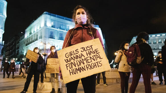 Eine junge Frau demonstriert auf der Straße zusammen mit anderen, sie hält ein Pappschild, auf dem geschrieben steht "Girls just want to have fun-damental human rights".