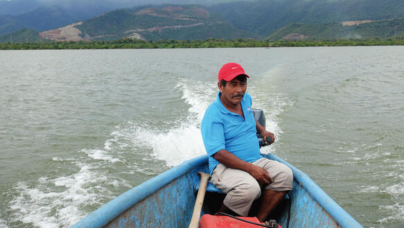 Ein Mann mit Schirmmütze sitzt auf einem Motorboot und steuert über einen See, im Hintergrund erheben sich Berge.
