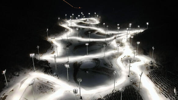Die Biathlonstrecke in China bei den Olympischen Spielen 2022 führt in engen Kurven durch die Landschaft, es ist Nacht, sie wird angestrahlt vom Licht zahlreicher Scheinwerfermasten, im Hintergrund steht die beleuchtete Skisprungschanze.