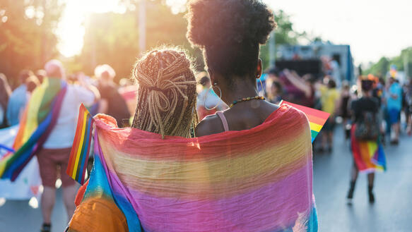 Rückenansicht von zwei jungen Frauen, die eine Regenbogenflagge um die Schultern haben und sich umarmen.