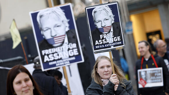 Das Bild zeigt zwei Personen, die demonstrieren. Sie haben Schilder mit dem Foto von Julian Assange in der Hand