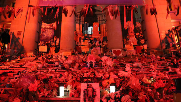 Das Bild zeigt Treppen vor einem Gebäude in roter Beleuchtung mit Kerzen, Bildern, Blumen und Spielsachen.