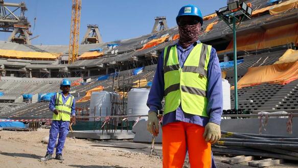 Zwei Menschen in Arbeitskleidung mit gelben Warnwesten auf einer Baustelle für ein Stadion. Ihre Gesichter sind verhüllt. 