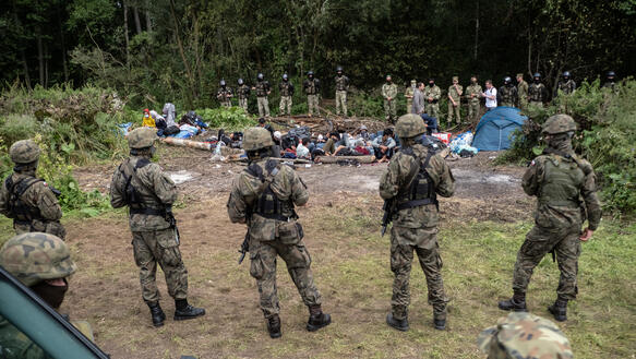 Das Bild zeigt mehrere Soldaten, die eine Gruppe von Menschen bewachen, die vor ihnen auf dem Boden kauern.