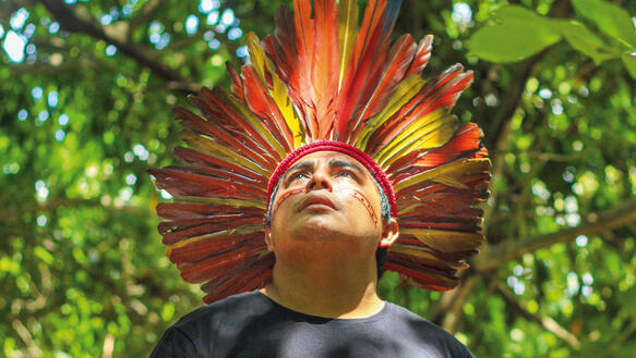 Ein brasilianischer indigener Aktivist steht in einem Wald, trägt T-Shirt und auf dem Kopf einen Federschmuck, er blickt nach oben.