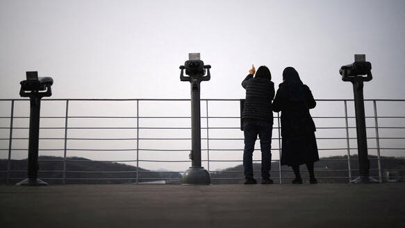 Zwei Frauen stehen an einem Geländer, hinter dem sich eine weite hügelige Landschaft erstreckt; öffentliche Ferngläser stehen vor dem Geländer.