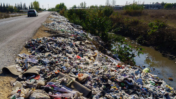 Die Böschung neben einer Landstraße, die zu einem kleinen Bach abfällt, ist voller Müll; auf der Straße fährt ein Auto.