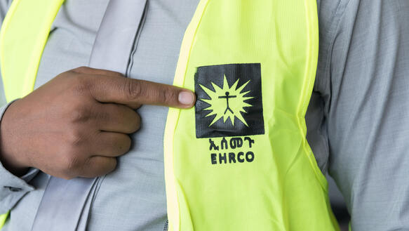 Der Oberkörper eines Menschen, der über einem blaugestreiften Hemd eine gelbe Warnweste mit einem Organisationslogo darauf trägt und mit dem Zeigefinger aufauf das Logo deutet. 