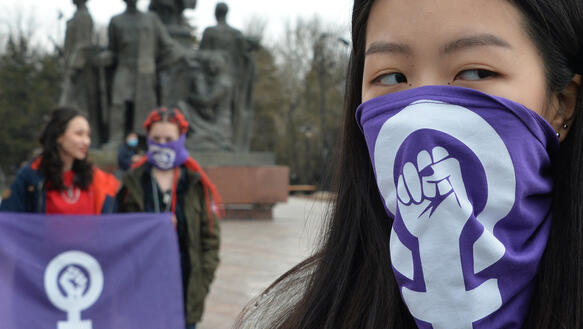 Eine junge Frau im Vordergrund trägt eine Maske mit einem Frauen*zeichen mit Faust in der Mitte. Im Hintergrund stehen zwei weitere Demonstrantinnen.