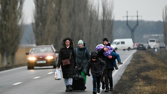Zwei Frauen mit drei Kindern laufen in Wintermantel mit Gepäck eine Straße entlang.