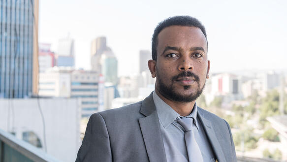 Ein äthiopischer Mann mit kurzen Haaren und kurzem Bart trägt ein Hemd und Jacket und Krwatte, steht vor der Skyline einer Großstadt, ernste Mimik. 