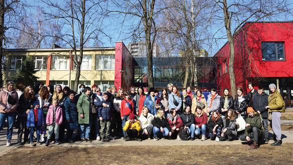 Kinder einer Schulklasse haben sich zusammen mit ihren Lehrerinnen zum Foto aufgestellt, hinter ihnen stehen Bäume und Schulgebäude.Schüler_innen und Lehrerinnen aus Charkiw vor ihrer neuen Schule in Vilnius