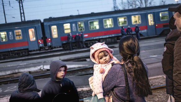 Eine Mutter mit Kindern steht auf einem Gleis am Bahnhof, im Hintergrund hält ein Zug, vor dem Menschen stehen.