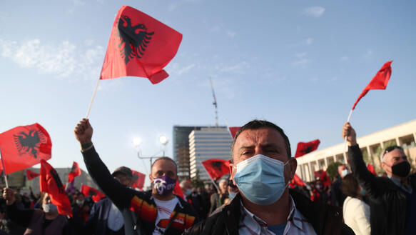 Viele Meschen mit Masken schwenken albanische Flaggen vor blauem Himmel.