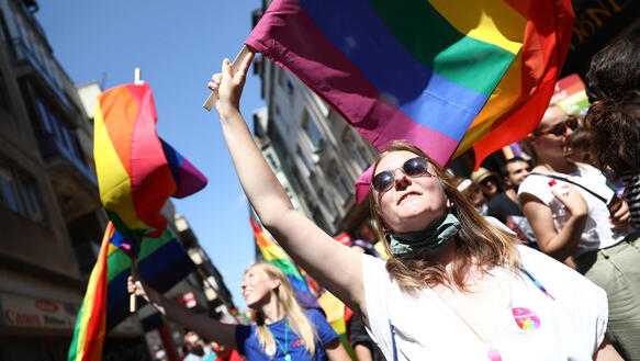 Zentral im Bild schwenkt eine junge Frau eine Regenbogenfahne. Im Hintergrund befinden sich weitere Demonstrant_innen.