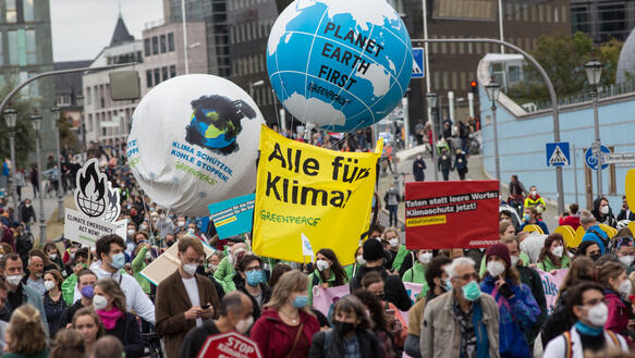 Eine Menschenmenge geht in eine Richtung, viele tragen FFP2-Masken, außerdem haben einige Plakate oder Ähnliches, auf denen beispielsweise steht: "Alle fürs Klima" oder "Planet Earth First". Im Hintergrund sind Hochhäuser zu sehen. 