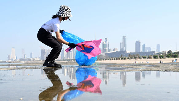 Ein Mädchen am Strand mit einer Mülltüte und Handschuhen. Im Hintergrund ist befindet sich die Skyline von Kuwait.