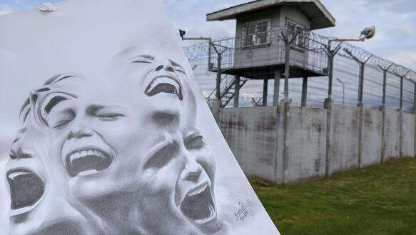 Das Bild zeigt eine Zeichnung im Vordergrund, im Hintergrund der Zaun und Wachturm einer Haftanstalt. Auf dem Bild sind verzweifelt schreiende Personen zu sehen.