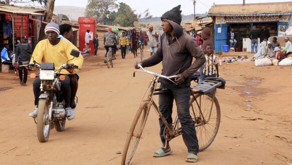 Ein Mann auf einem Fahrrad und ein Mann auf einem Motorrad auf einer staubigen Straße. Im Hintergrund sind weitere Personen und einige provisorisch aufgebaute Verkaufsstände.