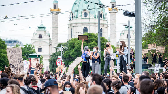 Foto einer Menschenmenge mit Schildern und Masken. Im Hintergrund befindet sich die Karlskirche.