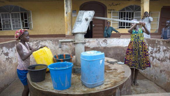 Ein Mädchen pumpt an einer Wasserpumpe, während ein weiteres Mädchen das Wasser in Eimer und Kanister füllt. Im Hintergrund befindet sich ein Gebäude.