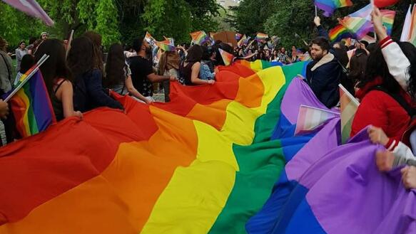 Das Bild zeigt eine Demonstration, die Personen tragen eine meterlange Regebogenflagge sowie kleine Fahnen