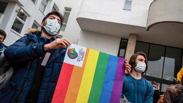 Zwei Männer mit Virenschutzmaske halten vor einem Gebäude eine Regenbogen-Fahne hoch.