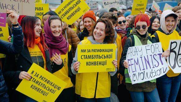 Das Foto zeigt eine große Menschenmenge. Im Vordergrund stehen mehrere junge Frauen, die Protestschilder hochhalten und sich anlächeln.