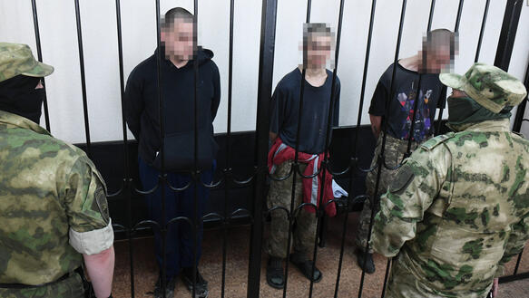 Drei Männer, die Gesichter unkenntlich gemacht, hinter Gitterstäben, davor zwei Männer in Tarnkleidung von hinten.