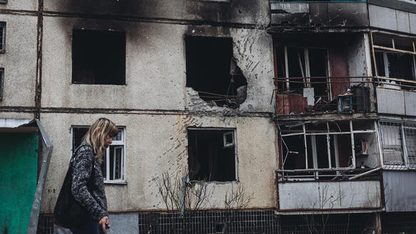 Das Bild zeigt eine Frau, die an einem Gebäude vorbeiläuft, das viele Einschusslöcher hat und Brandspuren
