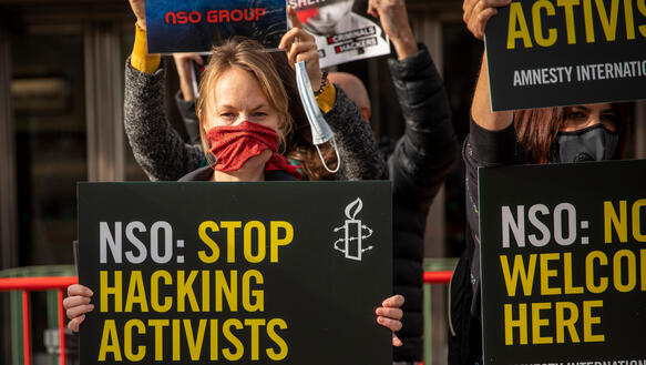 Eine Demonstration mit rotem Schal vor dem Mund hält ein Schild hoch, auf dem steht: „NSO: Stop Hacking Activists“.