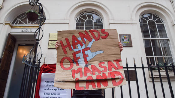 Das Bild zeigt eine Person, die vor einem Gebäude steht und ein Schild in der Hand hält: "Hands off Maasai Land"