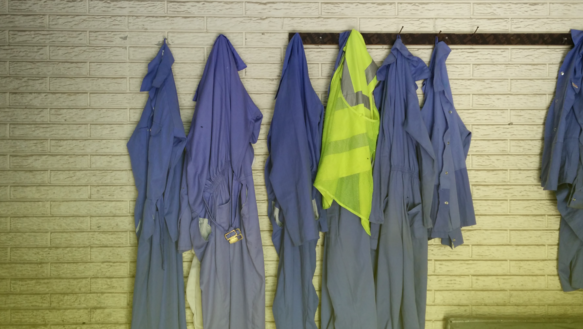 An einer Garderobenleiste und an Nägeln hängen an einer Wand blaue Arbeitsanzüge und eine gelbe Schutzweste.
