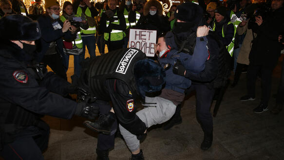 Ein Mann Frau hält ein Pappschild mit russischer Schrift in der Hand, während zwei Polizisten ihn an den Armen packen und wegzerren.