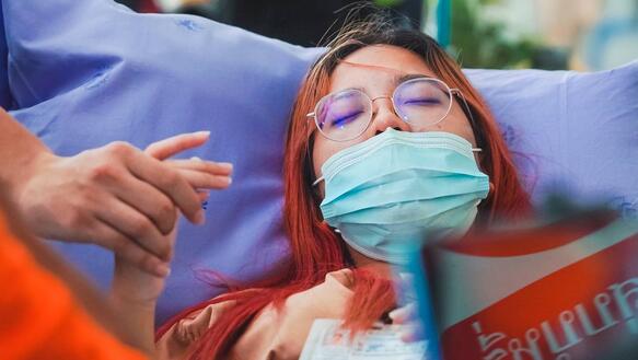 Das Bild zeigt eine Frau in einem Krankenbett mit geschlossenen Augen, jemand hält ihre Hand