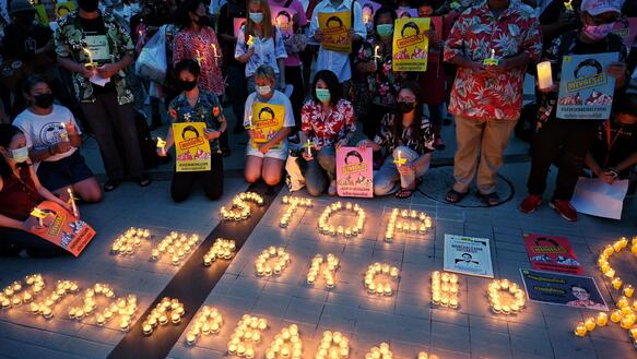 Das Bild zeigt eine Gruppe von Menschen mit Mundschutz bei einer Mahnwache. Im Vordergrund formen leuchtende Kerzen den Schriftzug "Stop enforced disappearances".
