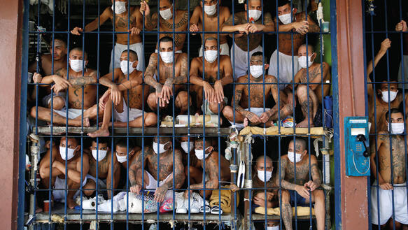 Männer tragen nichts als einen Mundnasenschutz und Boxershorts; sie sind hinter Gittern auf mehreren Ebenen übereinander eingepfercht.
