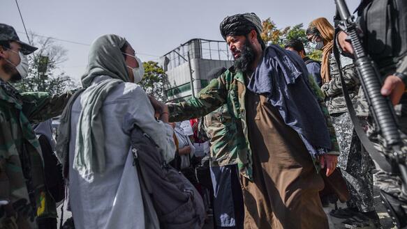 Ein Taliban-Kämpfer steht einer Frau gegenüber und hindert sie mit seinem Arm an ihrer Schulter am weitergehen. Um die beiden herum stehen weitere teils bewaffnete Männer.