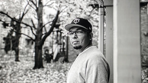 Ein Mann mit Sweat-T-Shirt und Baseball-Kappe trägt eine Brille und steht in einem Hauseingang, im Hintergrund stehen Bäume.