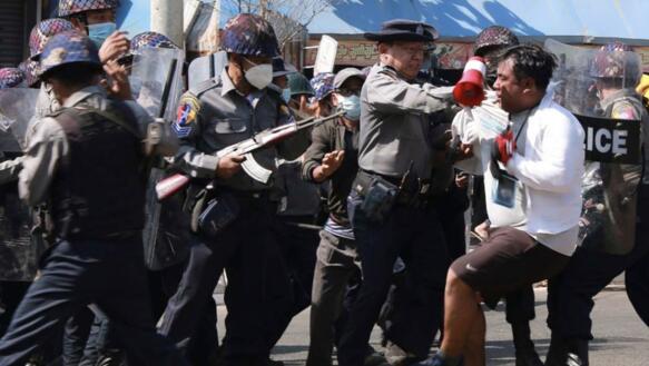 Ein Mann mit Megafon versucht, sich von einem Polizisten loszureißen. Um den Polizisten herum stehen viele weitere Polizisten, die Helme und Mundschutz tragen und teilweise mit Sturmgewehren bewaffnet sind oder Schutzschilde vor sich halten.