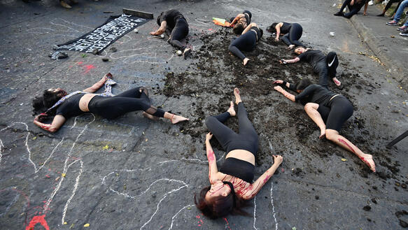 Frauen und Männer liegen mit roter Farbe auf den Gliedmaßen zwischen Erdklumpen und roter Farbe auf einer Straße und stellen sich tot, auf die Straße sind mit Kreide Körperumrisse gemalt, auch ein Transparent liegt dort.