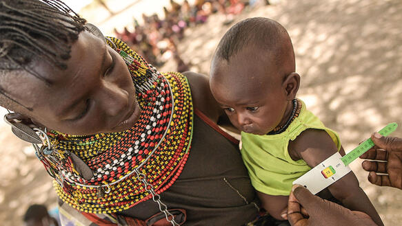 Eine Frau in Kenia trägt ihr Kind auf dem Arm, dessen Arm mit einem Maßband gemessen wird.