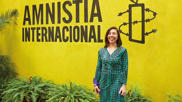 Eine mittelalte Frau mit halblangem Haar steht vor einem Banner mit der Aufschrift "Amnistía Internacional", sie lächelt und trägt ein langes Kleid; an ihrem rechten Handgelenk hängt ein Mundnasenschutz.