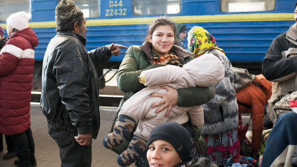 Menschen drängen sich auf einem Bahnsteig, im Hintergrund steht ein Zug, eine Mutter hält ihr Kleinkind auf den Armen und lächelt, alle Menschen tragen dicke Winterkleidung, manche Mützen oder Kopftuch.