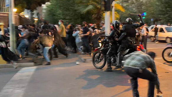 Das Bild zeigt zwei Polizisten auf einem Motorrad, die eine Menschenmenge vertreiben. Ein Polizist holt zum Schlag aus.