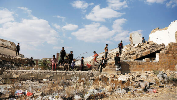 Jungen verschiedenen Alters sitzen und stehen in den Trümmern eines zerstörten Gebäudes, auch ein Mann steht dabei, aus den Trümmern ragen Eisenstangen.
