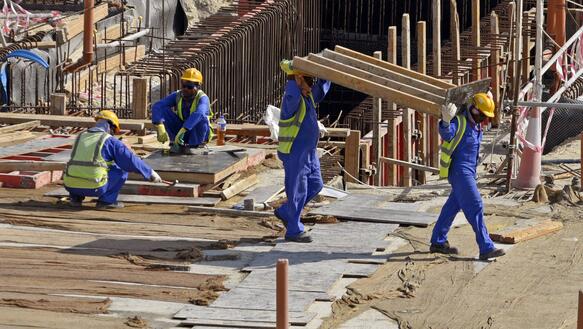 Das Bild zeigt eine Baustelle, mit mehreren Arbeitern, die gelbe Helme und Blaue Overalls tragen. 